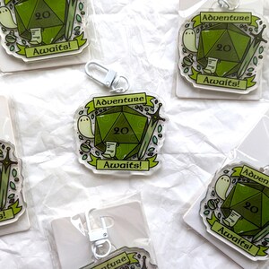 May include: Five green and white acrylic keychains featuring a 20-sided die, a ghost, and a sword. Each keychain has the text "Adventure Awaits!" printed on it.