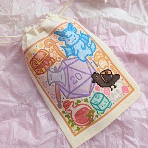 Könnte beinhalten: Eine weiße Kordeltasche mit einer bunten Illustration eines 20-seitigen Würfels, eines Drachen, einer Truhe, einer Krähe und anderen Fantasy-Elementen. Die Tasche ist perfekt zum Aufbewahren von Würfeln oder anderen kleinen Gegenständen.