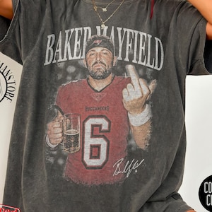 May include: A charcoal grey oversized t-shirt featuring a vintage-style graphic of Baker Mayfield in a red Buccaneers jersey with the number 6, holding a beer mug and gesturing with a middle finger. The text BAKER MAYFIELD is printed above.