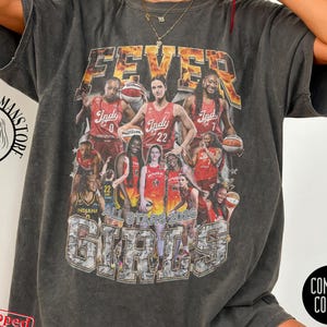 May include: A charcoal gray short-sleeve t-shirt featuring a graphic print of basketball players with the text FEVER and ALL STARS 2024 GIRLS. The shirt has a crew neckline and a relaxed fit. A circular logo and text are visible on the shirt.