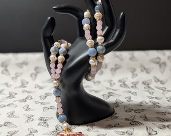 Puede incluir: Un collar de cuentas que alterna perlas de color azul claro, rosa pálido y blanco, acentuado con pequeños espaciadores dorados. El collar termina en un colgante de concha en forma de abanico con patrones de rayas marrones y blancas.