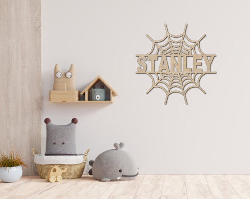 May include: A wooden spiderweb wall decor with the name "STANLEY" cut out in the center. The decor is hanging on a white wall in a nursery setting.