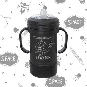 Może przedstawiać: Czarny kubek do picia z dwoma uchwytami, przezroczystą pokrywką i słomką. Kubek ma rysunek helikoptera i napis "BRAXTON" nadrukowany na nim. Tło jest białe z wzorem o tematyce kosmicznej.