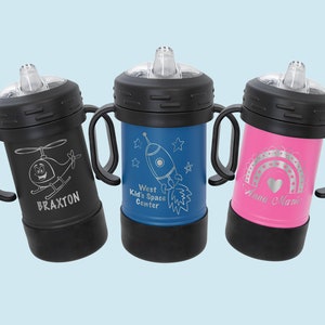 Może przedstawiać: Trzy stalowe kubki do picia z personalizowanymi grawerami. Kubki są czarne, niebieskie i r&oacute;żowe z czarnymi uchwytami. Grawery przedstawiają helikopter, rakietę i tęczę z sercem. Tekst na kubkach brzmi: "Braxton", "West Kids Space Center" i "Anna Marie".