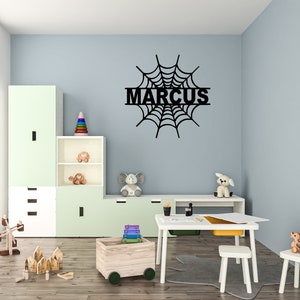 May include: Black wooden spiderweb wall decor with the name "MARCUS" in the center. The decor is hanging on a light blue wall in a child's room.