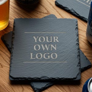 May include: Square slate coasters with the text "YOUR OWN LOGO" engraved in a light color. The coasters are dark gray and sit on a wooden surface, with glasses of beer and cans of beer in the background.