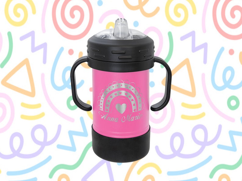 Może przedstawiać: R&oacute;żowy kubek ze stali nierdzewnej z czarną pokrywką i uchwytami. Kubek ma wz&oacute;r tęczy i serca z wygrawerowanym imieniem "Anna Marie".