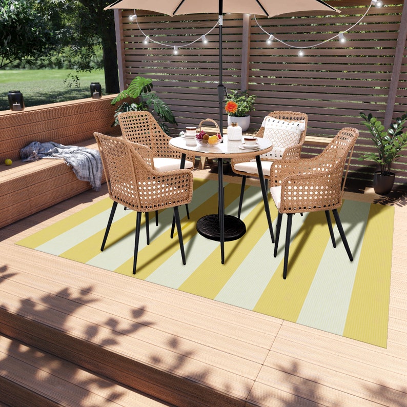 K&ouml;nnte beinhalten: Ein gelber und wei&szlig; gestreifter Outdoor-Teppich mit einem schwarzen Metalltisch und vier Korbst&uuml;hlen. Der Tisch hat eine wei&szlig;e Tischplatte und eine kleine Topfpflanze in der Mitte.