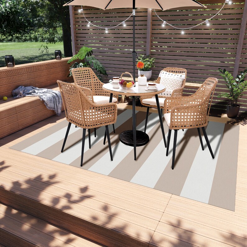 K&ouml;nnte beinhalten: Ein gestreifter Outdoor-Teppich mit einem braunen und wei&szlig;en Muster. Der Teppich liegt auf einem Holzdeck und verf&uuml;gt &uuml;ber einen Tisch und vier St&uuml;hle. Der Tisch ist rund und schwarz mit einer wei&szlig;en Tischplatte. Die St&uuml;hle sind aus Weide und haben schwarze Metallbeine.