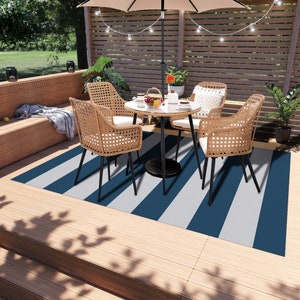 K&ouml;nnte beinhalten: Ein blauer und wei&szlig;er gestreifter Outdoor-Teppich mit einem Patio-Tisch und einem Stuhlset darauf. Der Tisch hat eine Glasplatte und die St&uuml;hle sind aus Weidengeflecht mit schwarzen Metallbeinen.
