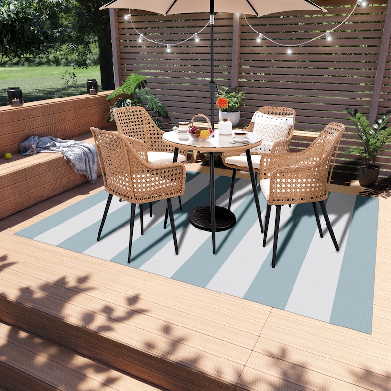 K&ouml;nnte beinhalten: Ein Patio-Set mit einem runden Tisch und vier St&uuml;hlen. Die St&uuml;hle sind aus Korbgeflecht und haben schwarze Metallbeine. Der Tisch ist ebenfalls aus schwarzem Metall und hat einen wei&szlig;en und blauen gestreiften Outdoor-Teppich darunter.