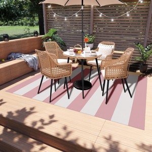 K&ouml;nnte beinhalten: Ein rosa-wei&szlig; gestreifter Outdoor-Teppich mit einem schwarzen Metalltisch und vier Korbst&uuml;hlen. Der Tisch hat eine Glasplatte und ist mit einer Blumenvase, Tellern und Tassen gedeckt.