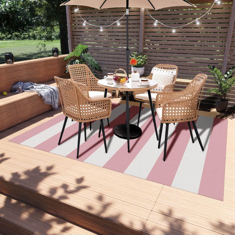 K&ouml;nnte beinhalten: Ein rosa-wei&szlig; gestreifter Outdoor-Teppich mit einem schwarzen Metalltisch und vier Korbst&uuml;hlen. Der Tisch hat eine Glasplatte und ist mit einer Blumenvase, Tellern und Tassen gedeckt.