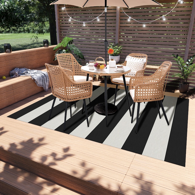 K&ouml;nnte beinhalten: Schwarz-wei&szlig;er gestreifter Outdoor-Teppich mit einem schwarzen Metalltisch und vier Korbst&uuml;hlen auf einem Holzdeck.