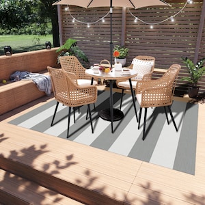 K&ouml;nnte beinhalten: Ein gestreifter Outdoor-Teppich in Grau und Wei&szlig; mit einem schwarzen Metalltisch und vier Korbst&uuml;hlen mit schwarzen Metallbeinen. Der Tisch hat eine Glasplatte und eine Schale mit Obst darauf. Die St&uuml;hle haben Kissen und sind um den Tisch herum angeordnet. Der Teppich liegt auf einem Holzdeck.