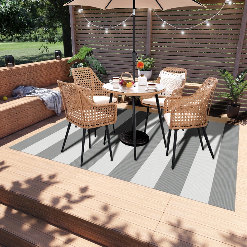 K&ouml;nnte beinhalten: Ein gestreifter Outdoor-Teppich in Grau und Wei&szlig; mit einem schwarzen Metalltisch und vier Korbst&uuml;hlen mit schwarzen Metallbeinen. Der Tisch hat eine Glasplatte und eine Schale mit Obst darauf. Die St&uuml;hle haben Kissen und sind um den Tisch herum angeordnet. Der Teppich liegt auf einem Holzdeck.
