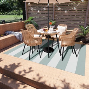 K&ouml;nnte beinhalten: Ein gestreifter Outdoor-Teppich in Gr&uuml;n- und Wei&szlig;t&ouml;nen, der auf einem Holzdeck platziert ist. Ein kleiner Tisch mit vier St&uuml;hlen steht auf dem Teppich. Die St&uuml;hle sind aus geflochtenem Weidengeflecht mit schwarzen Metallbeinen. Der Tisch ist rund und hat ein schwarzes Metallgestell.