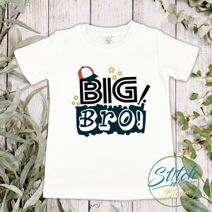 May include: White t-shirt with a black and blue graphic that says "BIG BRO!" with a red and white baseball cap above the text and gold stars around the graphic.
