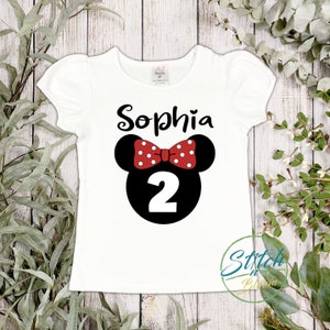 May include: White t-shirt with a black and white image of Minnie Mouse with a red bow and white polka dots. The number 2 is in the center of the image. The name Sophia is printed above the image.