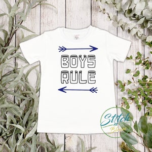 May include: White t-shirt with a black graphic that says "BOYS RULE" with blue arrows on either side of the text.