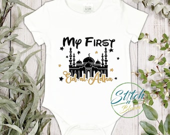 May include: White baby onesie with black text that reads "My First Eid al-Adha" with a black silhouette of a mosque and gold stars.