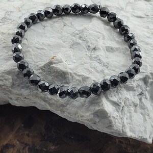 Peut inclure: Un bracelet de perles noires avec des perles &agrave; facettes. Le bracelet est pr&eacute;sent&eacute; sur une surface de pierre grise clair et marron. Les perles ont un aspect brillant et r&eacute;fl&eacute;chissant.