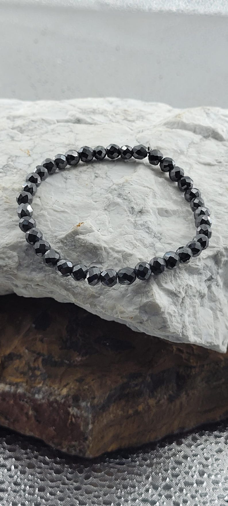 Peut inclure: Un bracelet de perles noires avec des perles &agrave; facettes. Le bracelet est pr&eacute;sent&eacute; sur une surface de pierre grise clair et marron. Les perles ont un aspect brillant et r&eacute;fl&eacute;chissant.