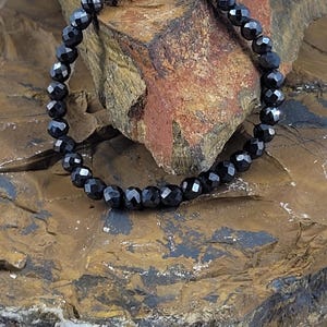 Peut inclure: Un bracelet de perles noires avec des perles &agrave; facettes. Le bracelet est pr&eacute;sent&eacute; sur une surface de pierre textur&eacute;e, marron et beige. Les perles refl&egrave;tent la lumi&egrave;re, cr&eacute;ant un effet scintillant.