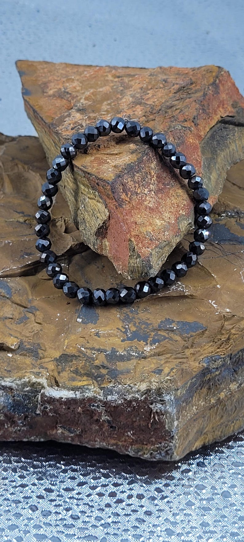 Peut inclure: Un bracelet de perles noires avec des perles &agrave; facettes. Le bracelet est pr&eacute;sent&eacute; sur une surface de pierre textur&eacute;e, marron et beige. Les perles refl&egrave;tent la lumi&egrave;re, cr&eacute;ant un effet scintillant.