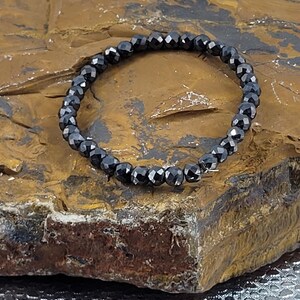 Peut inclure: Un bracelet de perles noires avec des perles &agrave; facettes. Le bracelet est compos&eacute; de petites perles noires rondes enfil&eacute;es ensemble. Le bracelet est pr&eacute;sent&eacute; sur une surface brune textur&eacute;e.