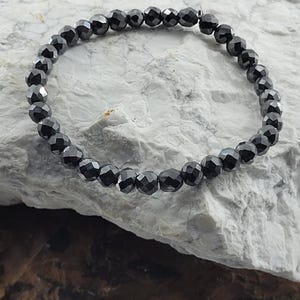 Peut inclure: Un bracelet de perles noires avec des perles &agrave; facettes. Le bracelet est enfil&eacute; sur un cordon souple et repose sur une surface de pierre textur&eacute;e et claire. Les perles ont une qualit&eacute; r&eacute;fl&eacute;chissante.