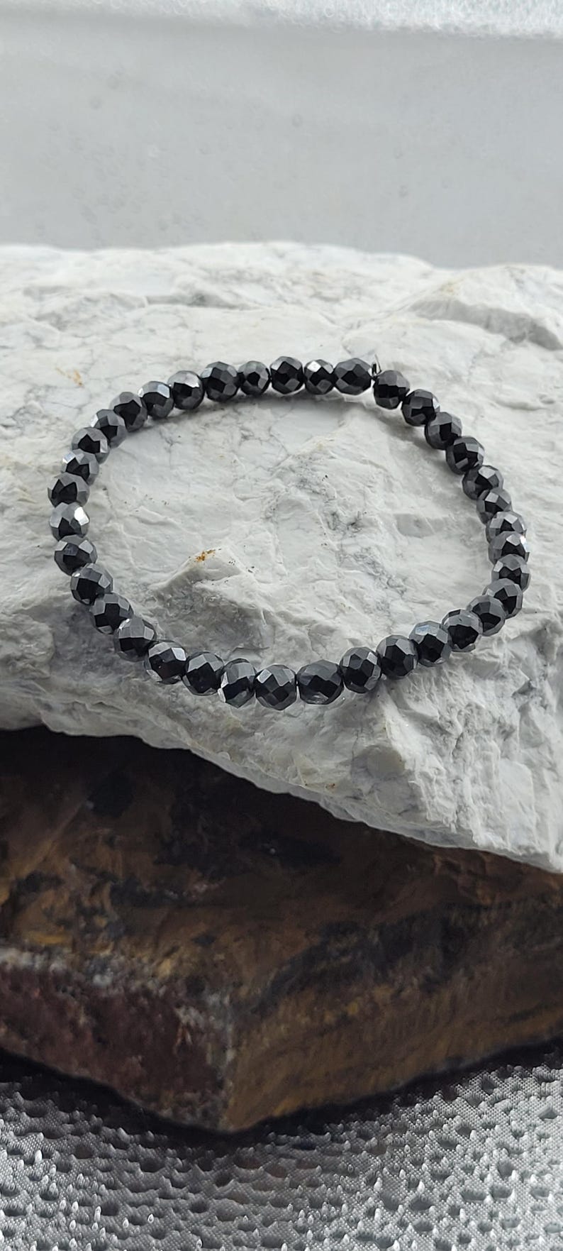 Peut inclure: Un bracelet de perles noires avec des perles &agrave; facettes. Le bracelet est enfil&eacute; sur un cordon souple et repose sur une surface de pierre textur&eacute;e et claire. Les perles ont une qualit&eacute; r&eacute;fl&eacute;chissante.