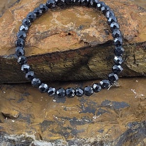 Peut inclure: Un bracelet de perles noires avec des perles &agrave; facettes. Le bracelet est mont&eacute; sur un cordon &eacute;lastique et est pr&eacute;sent&eacute; sur une surface brune textur&eacute;e. Les perles ont une qualit&eacute; r&eacute;fl&eacute;chissante.
