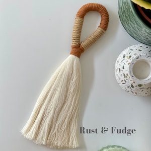 Peut inclure: Un pompon blanc avec une boucle en corde marron et beige. Le pompon est fait de fil doux et moelleux et a une longue frange fluide. La boucle de corde est nouée en haut du pompon. Le texte "Rust & Fudge" est visible dans le coin inférieur droit de l'image.
