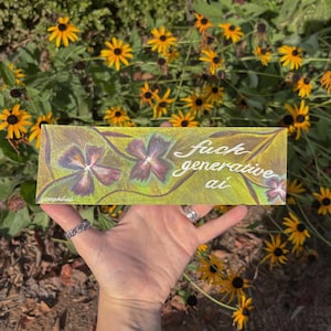 May include: A rectangular art piece with a yellow and green background featuring hand-drawn purple flowers and the white cursive text "fuck generative ai" written across the center, held against a backdrop of yellow coneflower blossoms.