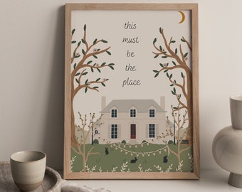 以下が含まれることがあります： 「this must be the place」のテキストが入った家のイラストが描かれた、額入りのアートプリント。アートワークには、木、三日月、装飾的なライトが含まれており、すべてソフトでニュートラルなカラーパレットで表現されています。フレームは明るい色の木材で作られています。