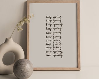 以下が含まれることがあります： 「keep going」の文字が繰り返されるフレーム入りのプリント。明るい木製のフレームに入っており、装飾的な花瓶と石の球体と一緒に展示されています。