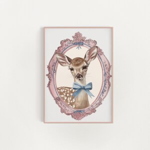 Könnte beinhalten: Ein gerahmter Aquarelldruck mit einem Reh mit einer blauen Schleife, eingebettet in einen rosa-blauen, verzierten ovalen Rahmen. Das Reh hat braunes Fell mit weißen Flecken und große Ohren. Der Rahmen ist detailliert.