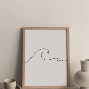 May include: A framed art print featuring a minimalist line drawing of a wave in black against a light gray background. The artwork is displayed in a natural wood frame. The image is set on a white surface with a neutral background.