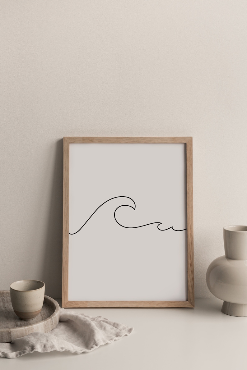 May include: A framed art print featuring a minimalist line drawing of a wave in black against a light gray background. The artwork is displayed in a natural wood frame. The image is set on a white surface with a neutral background.
