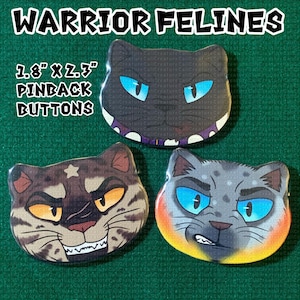 May include: Three cat-shaped pinback buttons labelled Warrior Felines, measuring 4.57 cm by 5.84 cm. One button features a black cat with blue eyes, one a brown tabby with yellow eyes, and one a grey spotted cat with bright blue eyes.