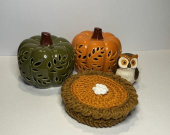 May include: An autumn-themed still life featuring a green and orange ceramic pumpkin, a small owl figurine, and a crocheted pie. The pumpkins have leaf-shaped cutouts. The pie is brown and orange with a white dollop.