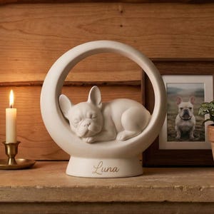 May include: A white ceramic sculpture featuring a sleeping French bulldog puppy nestled inside a circular frame. The base of the sculpture is inscribed with the name Luna in a cursive script. The object is displayed on a stone mantelpiece.
