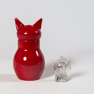 May include: A red ceramic cat-shaped container with a clear glass snow globe ornament. The snow globe has a gold-colored ornament inside.