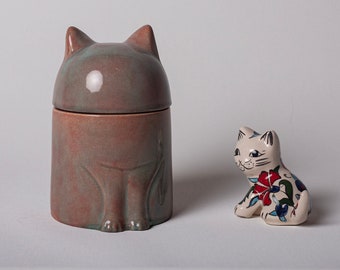 Op de afbeelding: Een keramische kattenvormige container met een deksel, geschilderd in tinten bruin en groen. Een kleiner keramisch kattenbeeldje met een witte basis en kleurrijke bloemmotieven staat op een wit oppervlak.