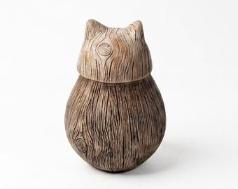 May include: A small, rounded, cat-shaped figurine crafted with a visible wood grain texture. The object features a light brown, smooth upper section representing the head and a darker, vertically textured lower body with natural wood patterns.