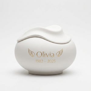 Pode incluir: Uma urna de cer&acirc;mica branca com corpo arredondado e tampa. A urna &eacute; decorada com asas douradas e o nome "Olivia" juntamente com os anos 1982-2025. A urna tem um acabamento fosco e uma textura suave.