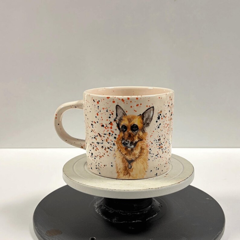 A ceramic mug showing a German Shepherd painting and paint splatters rests on a rotating stand.
