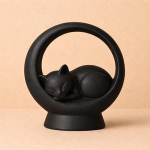 Puede incluir: Figura de gato negro de cerámica durmiendo dentro de un soporte en forma de media luna. El gato está acurrucado, con los ojos cerrados. El soporte tiene un diseño circular y se asienta sobre una base redonda. Es un objeto decorativo.