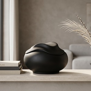 Pode incluir: Um pote decorativo preto fosco com um design de tampa org&acirc;nico &uacute;nico. O pote est&aacute; sobre uma superf&iacute;cie clara, com uma pilha de livros e plantas secas ao fundo. A forma do pote &eacute; arredondada com uma superf&iacute;cie texturizada.